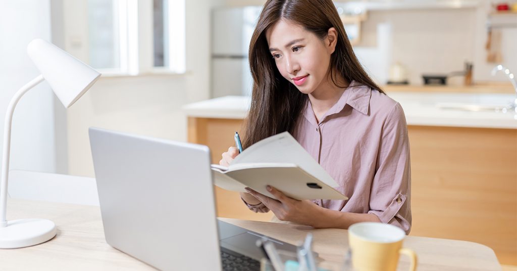 Jovem mulher estudando em frente ao notebook e um livro nas mãos